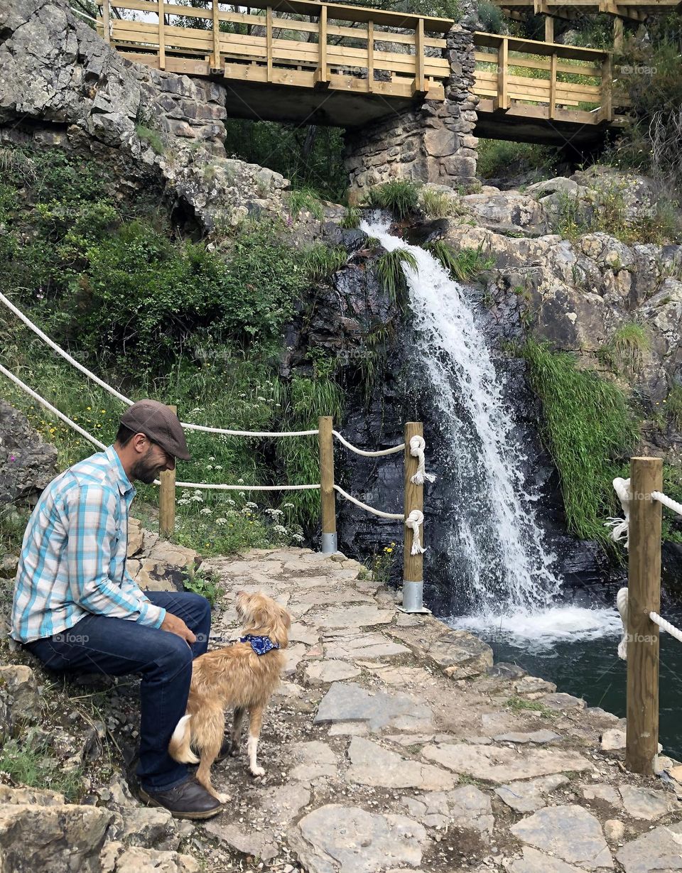 A man & his dog pause on a walk to enjoy the waterfall and scenery 