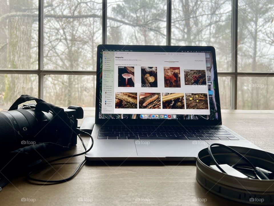 Photography workspace on wooden drafting table. Camera is plugged into laptop and a metal round container holds equipment