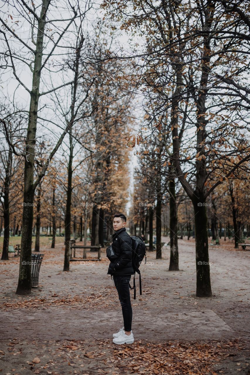 A young boy is feeling the fall vibes by standing in the middle of park with trees and brown leaves 