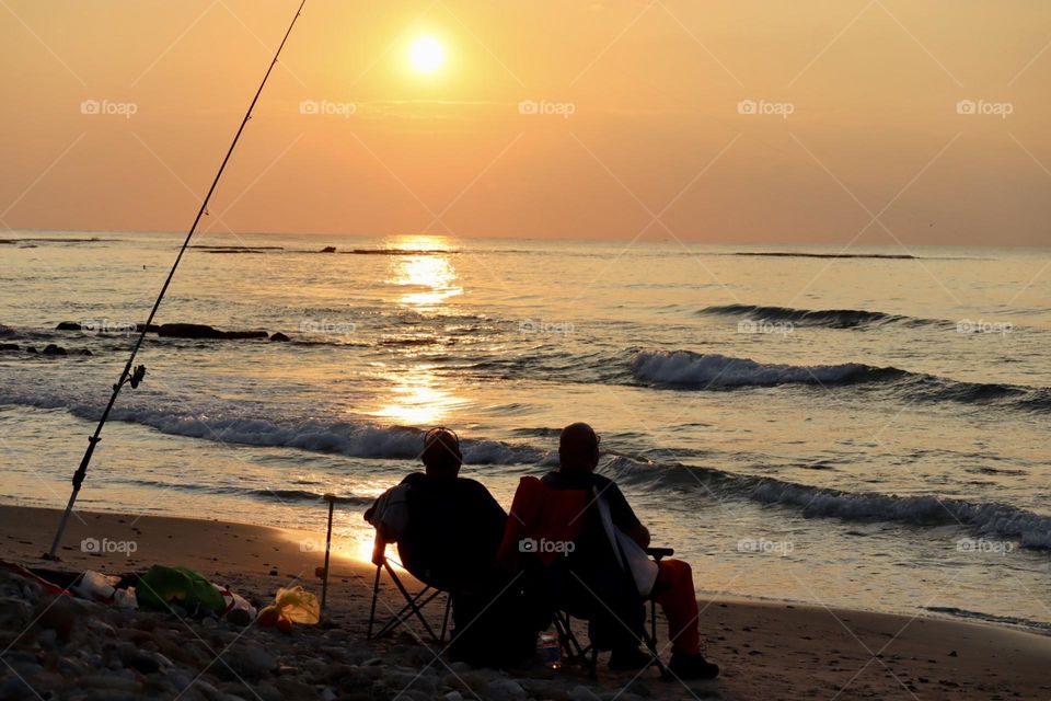 Two men sitting on the beach watching sunset after day of fishing together 