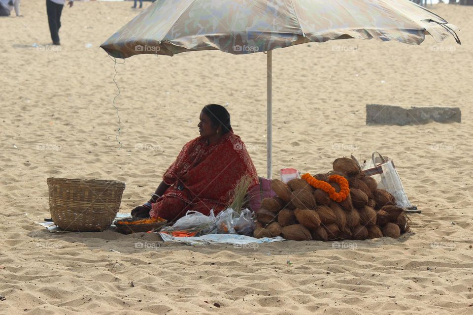 This picture describes a coconut sealing woman who is sitting under the shade of an umbrella over the beach.