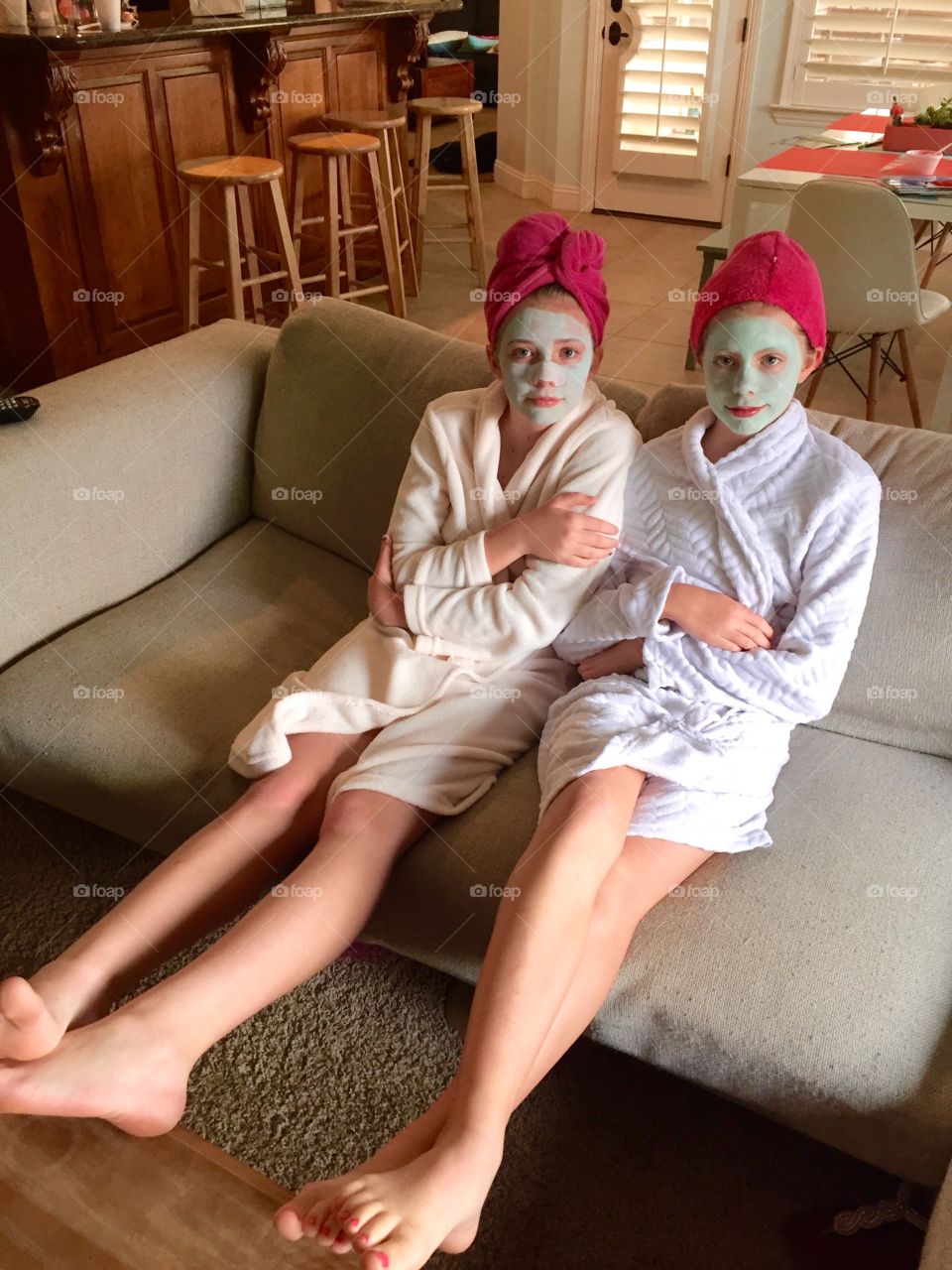 Spa day. Two girls in robes and facial masks sit next to each other on a couch.
