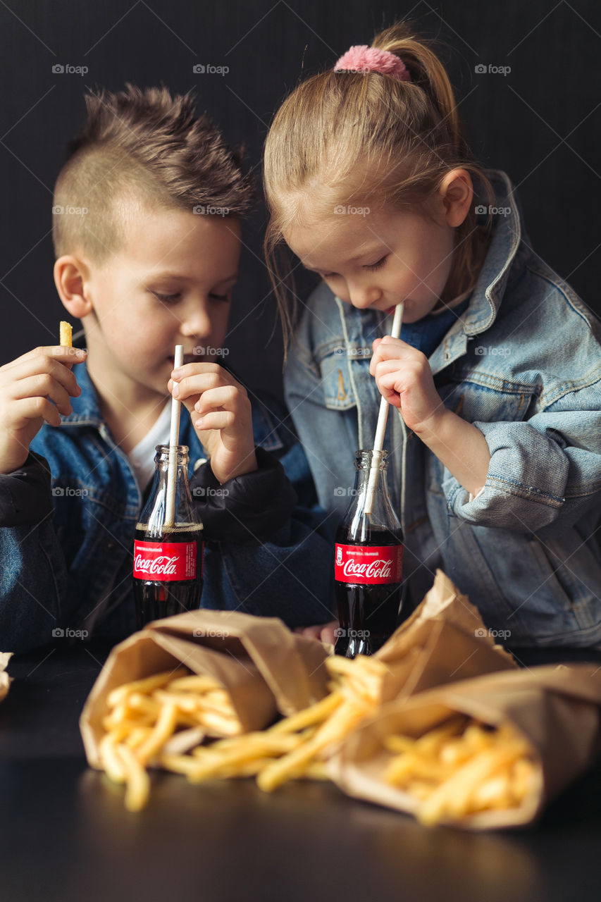 stylish children, dressed in denim clothes, drinking Coca-Cola.  They eat, relax, communicate at home.  sister and brother drink coca cola