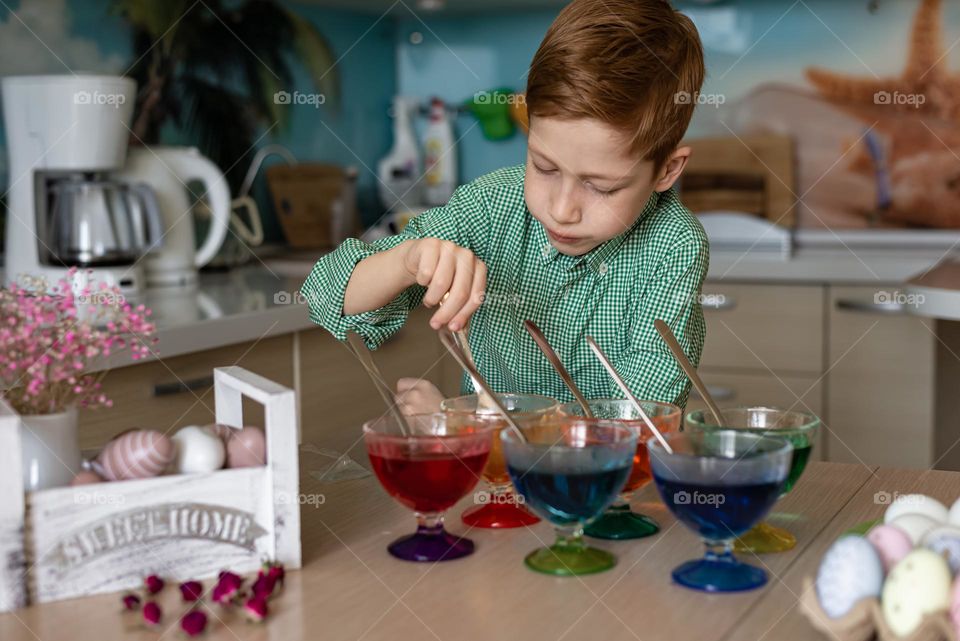 Child red-haired boy in a green shirt paints Easter eggs at home in the kitchen, craft product