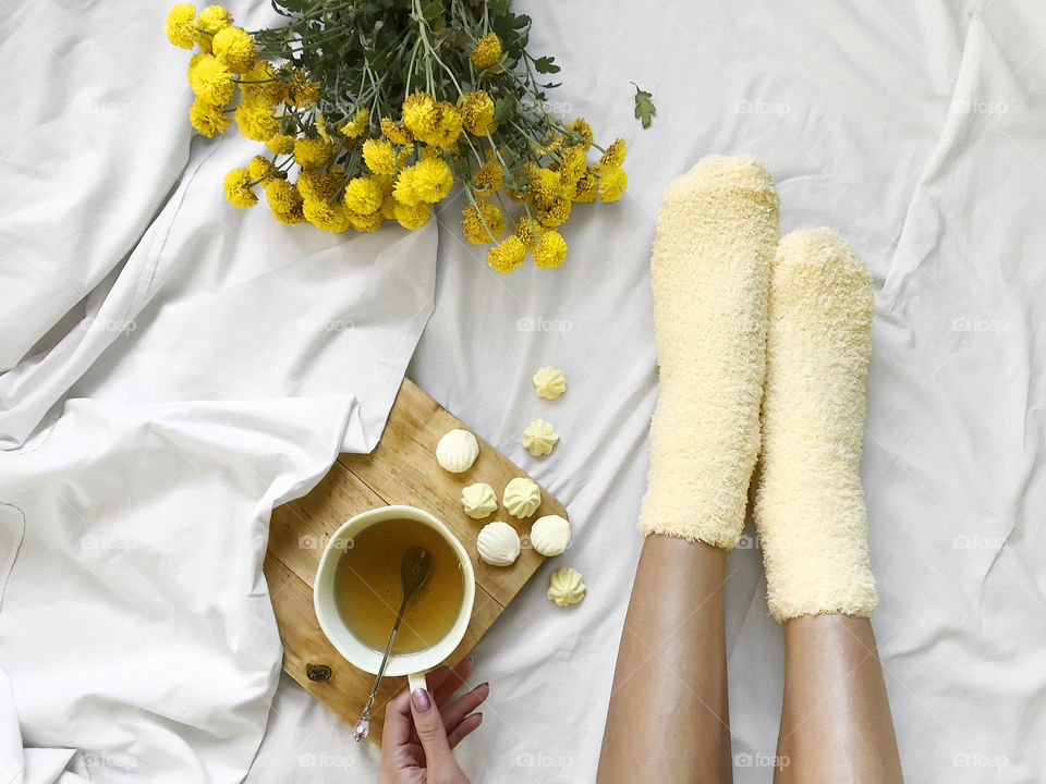 Young woman wearing yellow cozy socks and enjoying her cup of tea in bed with yellow bouquet of spring flowers 