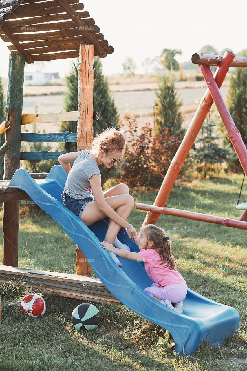 Teenage girl playing with her younger sister in a home playground in a backyard. Happy smiling sisters having fun on a slide together on summer day. Real people, authentic situations