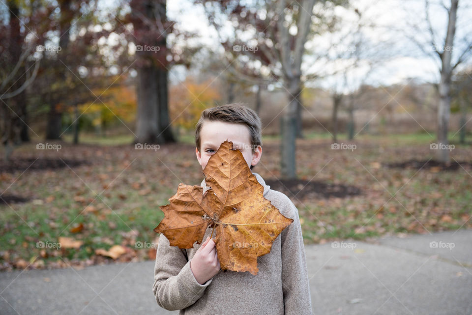 Young boy standing outdoors in autumn and covering his face with a large fall leaf 