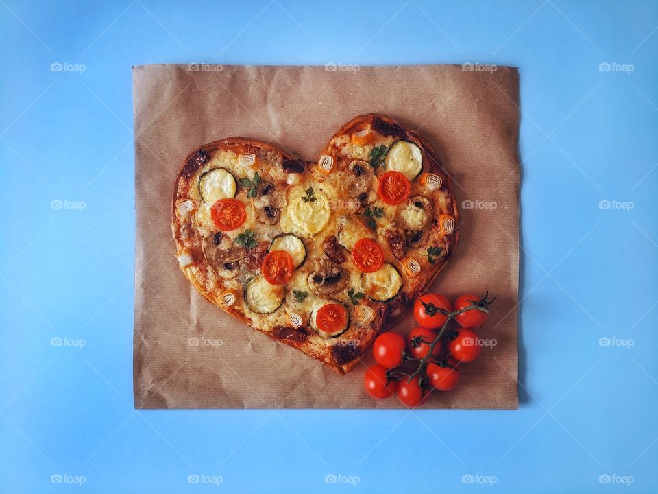 Delicious vegetarian pizza in the shape of a heart for Valentine's Day with tomatoes, vegetables and cheese on a blue background.