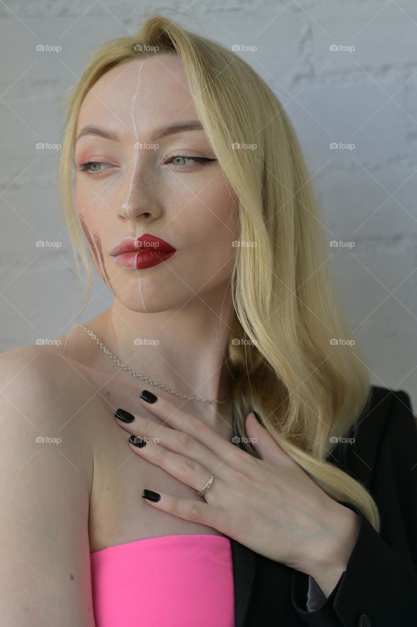 Close-up portrait of a blonde serious girl with different make-up on half of her face with her hand on the chest on white background