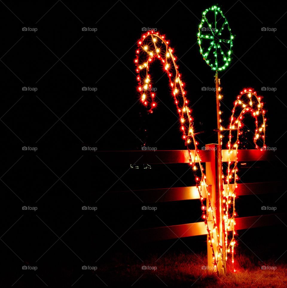 Christmas lights shaped like large candy canes and a lollipop against a wooden fence lit up a dark at night. 
