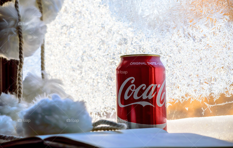 Closeup of red coke can on table with open book and cozy pompons near sunny window covered in frost crystals fun winter refreshment 