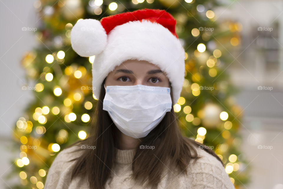 Beautiful woman in a red Santa Claus hat and a protective medical mask stands against the background of a decorated Christmas tree.  Concept: celebrating merry christmas during the coronavirus epidemic social distance