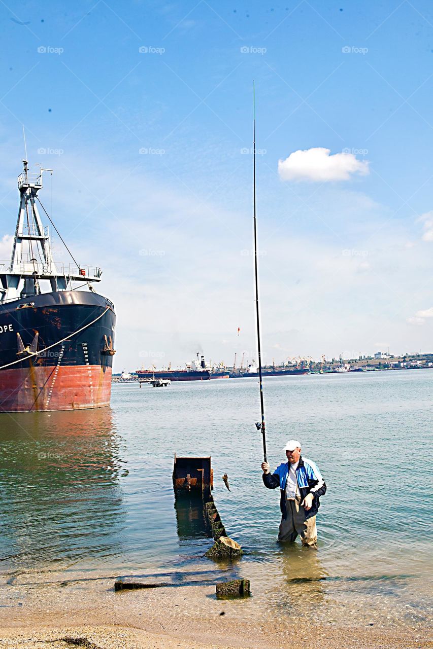 A man catches fish in the sea near a cargo ship