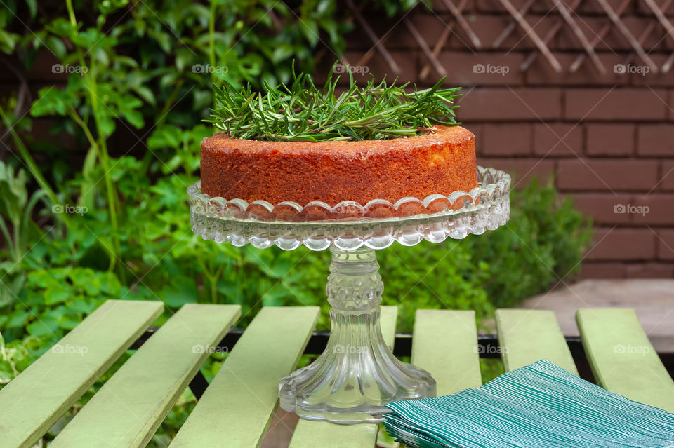 A perfect homemade sponge cake on crystal cake stand on green garden table decorated with rosemary herb.