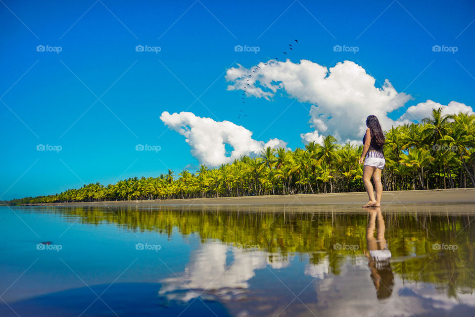 Composition of the sea water reflection of a girl observing the beautiful landscape of green palm trees contrasted by the blue sky