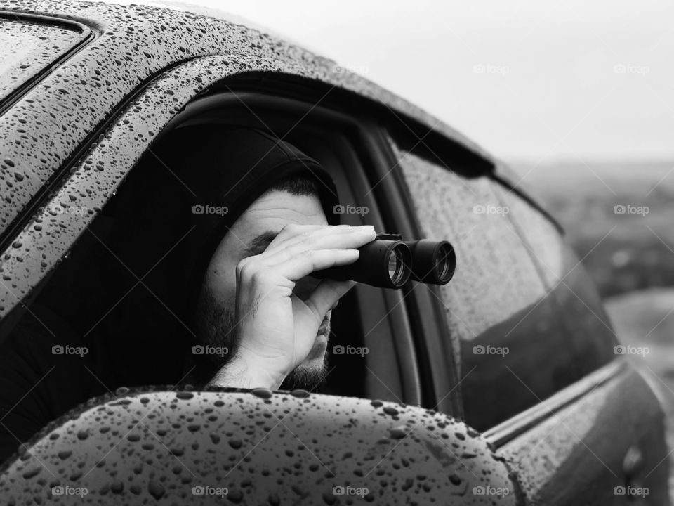 Young man watching through the binoculars out of car during the rainy autumn day 