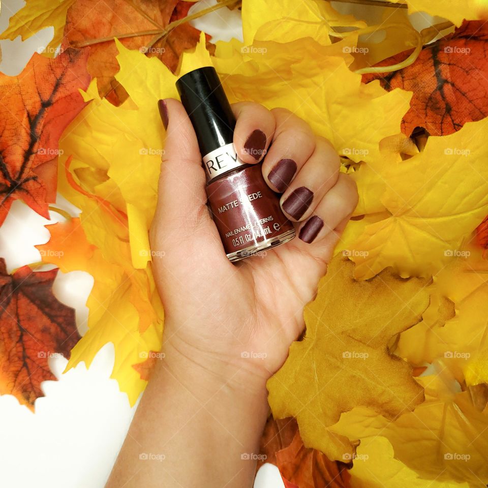 Holding a fall color matte suede nail polish with fall colored leaves in the background on a white surface.