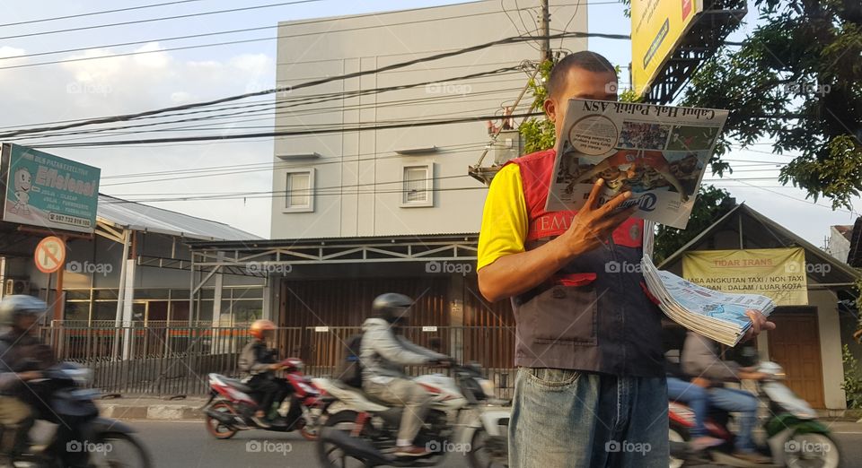 A newspaper seller reads a copy of the Tribun Jogya by the side of the road, Seville while early morning commuters stream past in mopeds, in #Yogyakarta, Indonesia