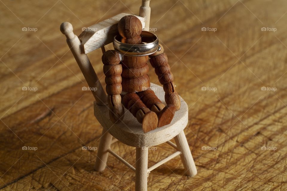 A wooden man used as an artist drawing tool sits in a miniature toy chair on a cutting board with a gold ring around its neck.