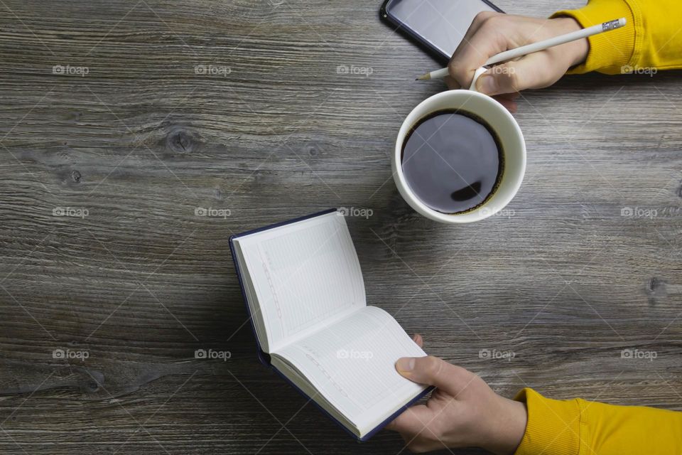 A man drinks coffee in the morning, works on the phone and makes notes in pencil in a notebook. Men's hands hold a white cup of coffee and a mobile phone in a yellow jacket. The hands lie on a gray wood background.