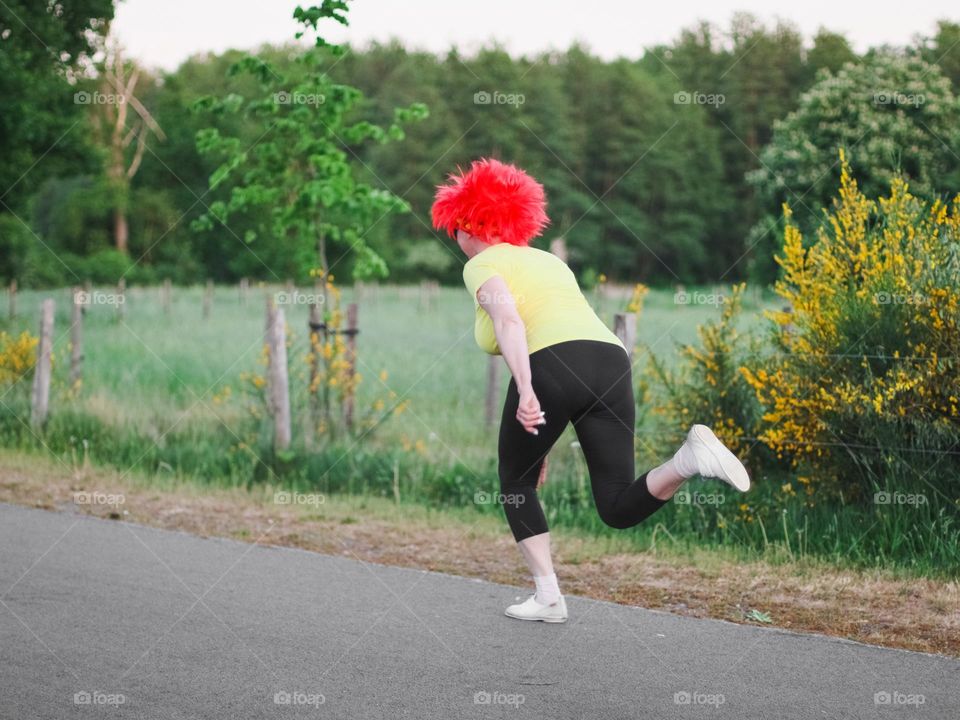 Portrait of a middle-aged caucasian woman in a yellow t-shirt, black leggings and a red wig from the back runs along a village road against a blurred field. Belgium day concept.