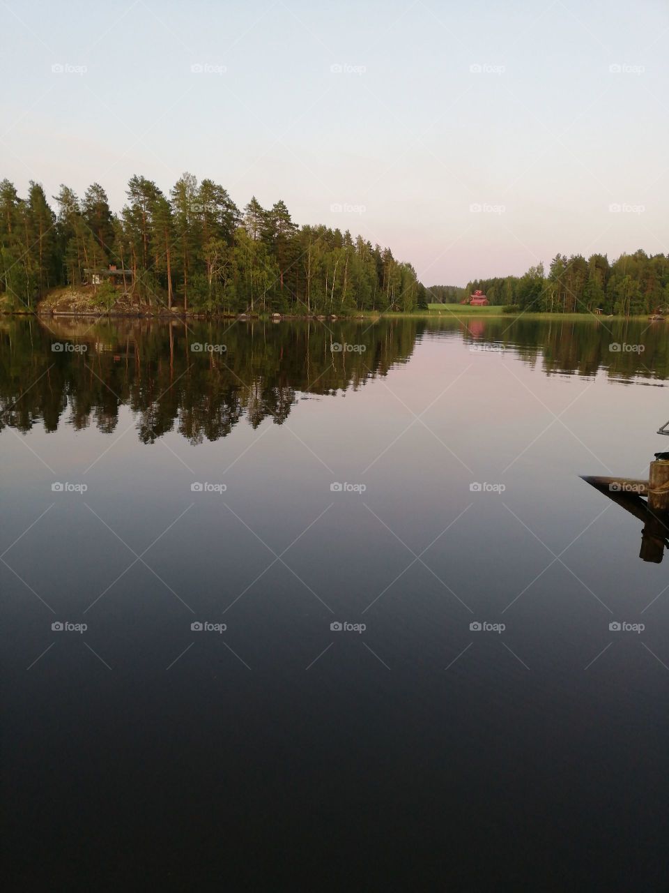 In a summer evening a little bit rippling, calm water in the front, on the other side of the lake are a lake side sauna, a summer cottage, a farmhouse, a green field and woods. No clouds in the sky.