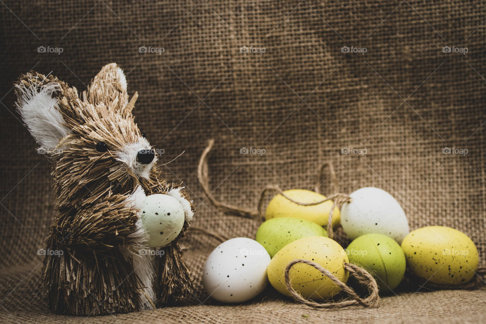straw Easter bunny, with a pile of colorful decorative Easter eggs, set against a rustic brown background