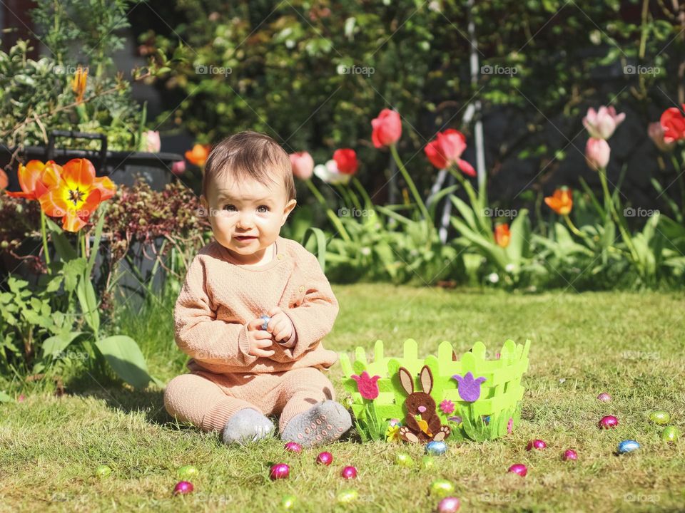 A beautiful caucasian little girl sits on the lawn with a felt basket and chocolate Easter eggs scattered around in shiny wrappers and holds a candy in her hands while smiling at the camera on a clear sunny spring day, close-up side view. Easter trad