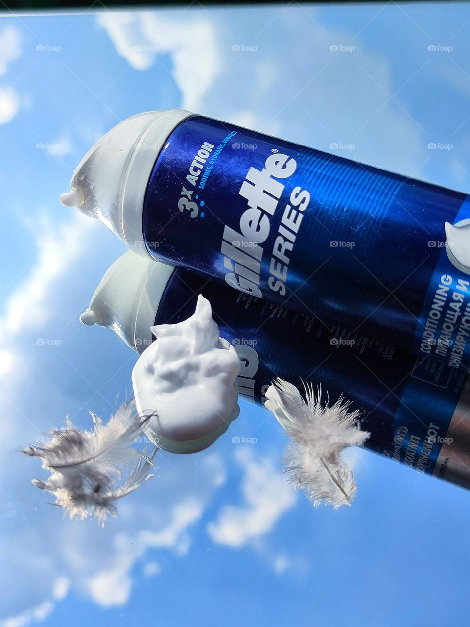 Shaving foam "Gillette" on a mirror surface. Nearby is white foam and two feathers, which are reflected in a mirrored surface with a blue sky and white clouds.