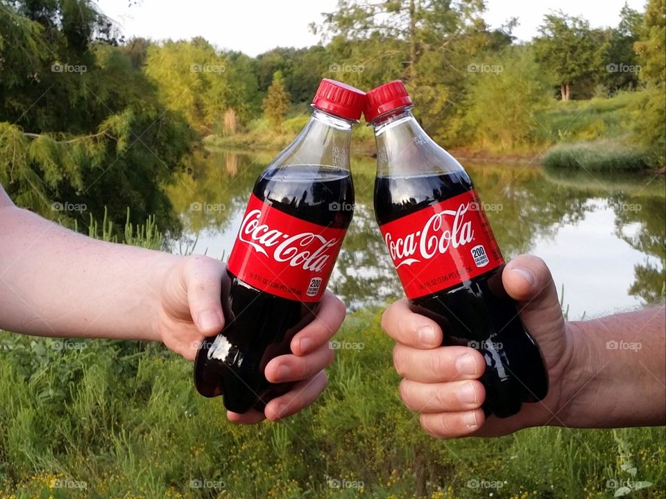 Two men holding Coca cola bottles in front of a pond with trees reflected in the water...Cheers. .liquids are cool