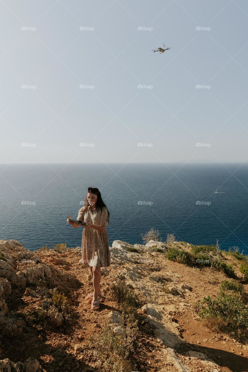 One beautiful young brunette girl controls a drone filming herself, standing on a high mountain against the sky on a clear sunny summer day, close-up side view.