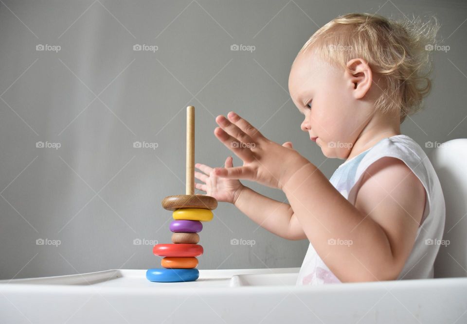 Little baby girl sitting in feeding chair and playing with wooden pyramid bricks