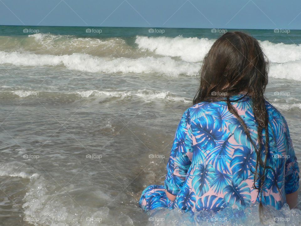 Glimmer moment at the beach with a girl child dressed with blue leaves flowers motifs designed on modest swim dress suit with the young girl sitting on the shore with long hair and loving the waves and warm ocean water.