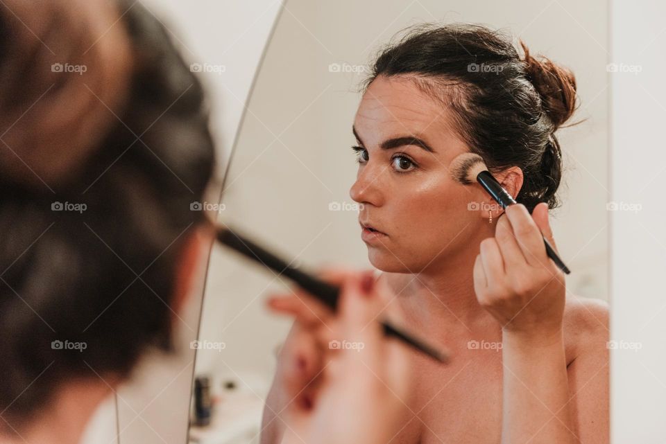 Mirror photo of toung woman using makeu-up brush to apply bronzer