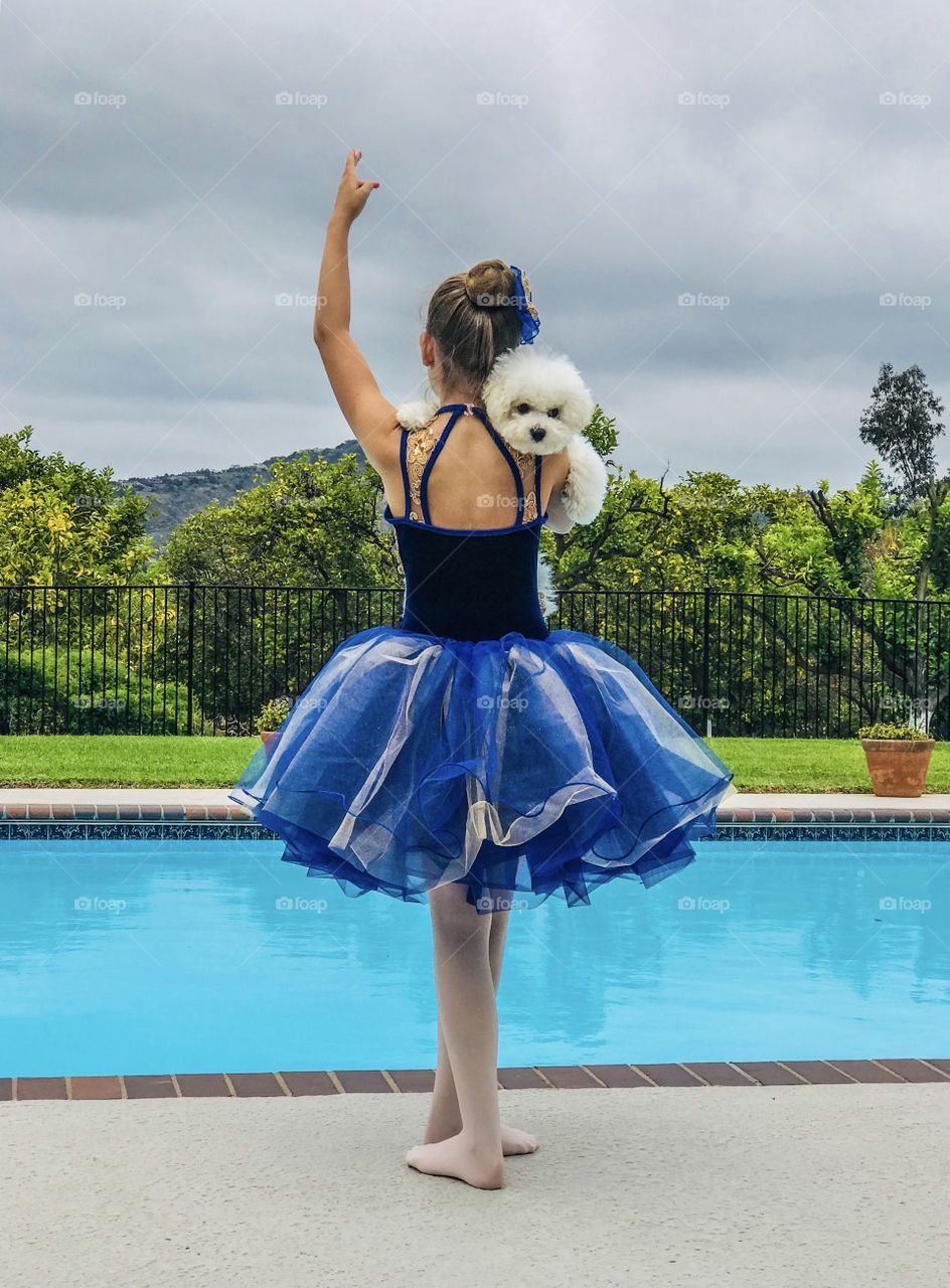 A ballerina in a costume of blue tulle and velvet strikes a pose while holding a furry puppy dog poolside with grove and mountain the background with an overcast sky. 