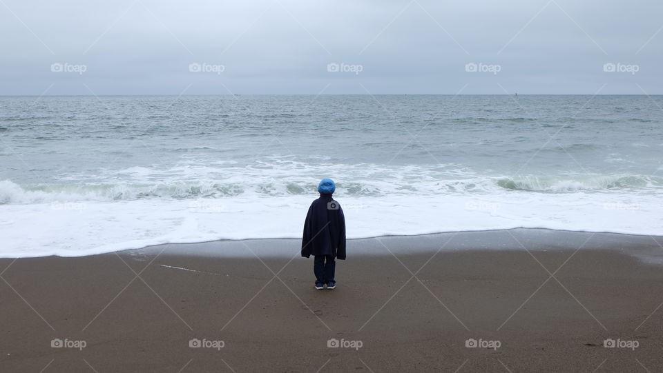 Boy wearing dad's oversized jacket and stares at the incoming waves.