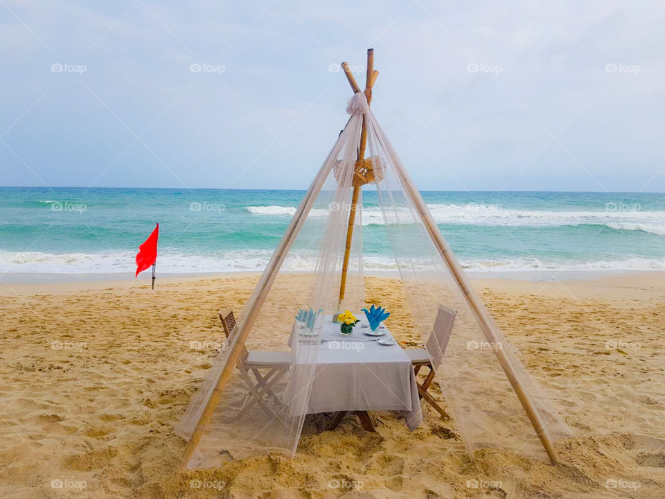 Sandy beach on a tropical island.  Served table for two under a transparent fabric canopy on the bank of a turbulent ocean with waves.  Romantic date on the ocean