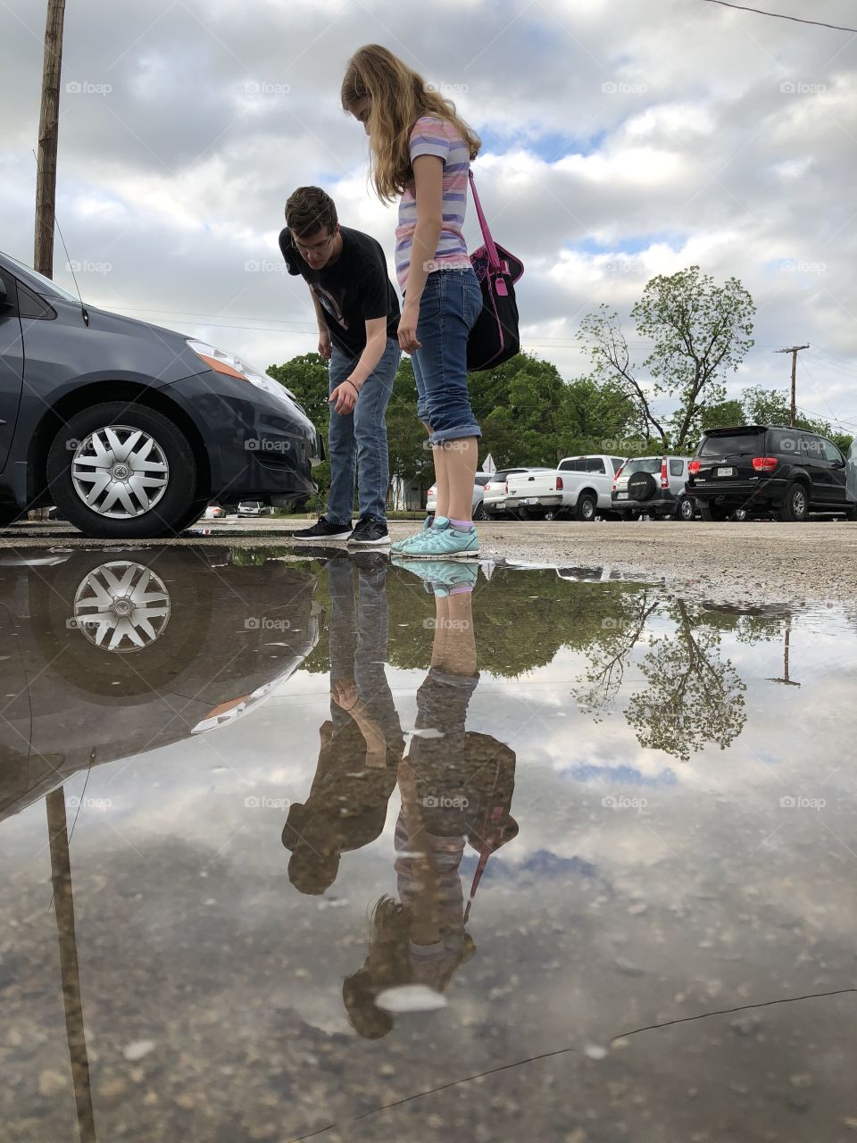 Brother and sister are reflected in puddle as they lean over it with bright blue sky and clouds and cars in background 