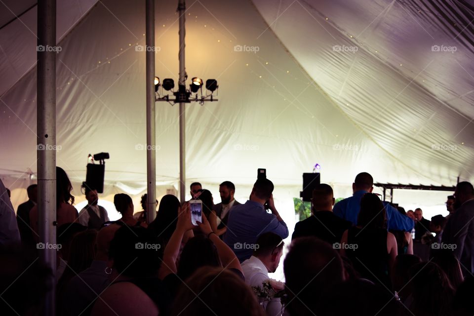 People holding phones and cameras inside a party tent at a wedding reception to take a photo of the bride and groom’s first dance