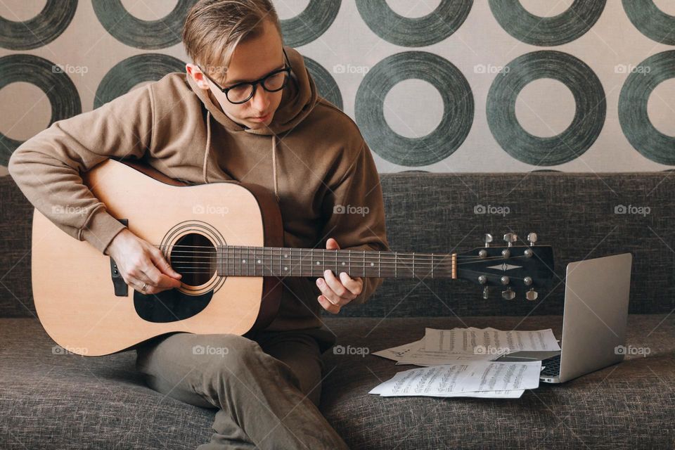 Young millennial man playing guitar learning online using music sheets 