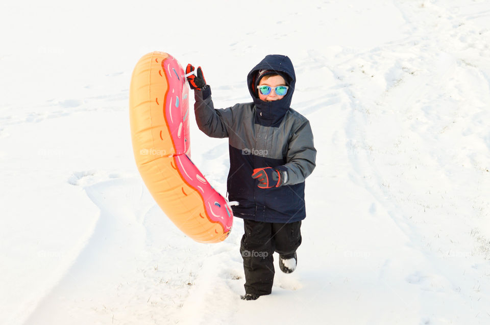 Young boy happily running in the snow outdoors with a donut inner tube to go sledding outdoors