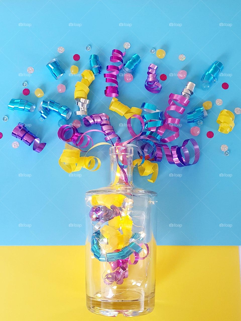 Colour explosion flatlay on a blue and yellow background composed of pink, purple, blue, silver and yellow gift package ribbons placed as if explosing out of a clear bottle