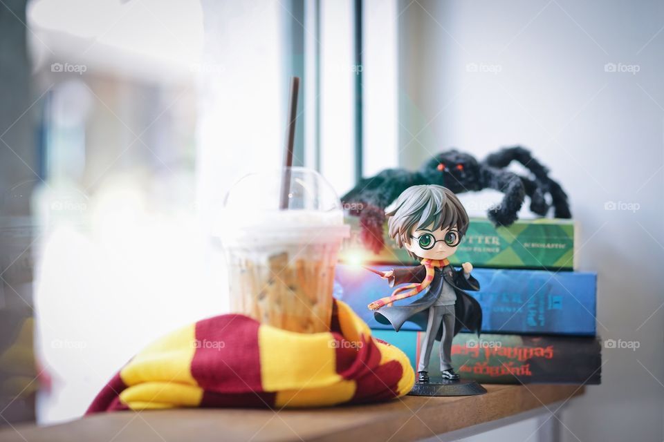 Bangkok, Thailand - Oct 13, 2019 : A photo of Harry Potter holding magical wand with a cup of iced coffee wrapped in Gryffindor scarf with rows of Harry Potter books series.