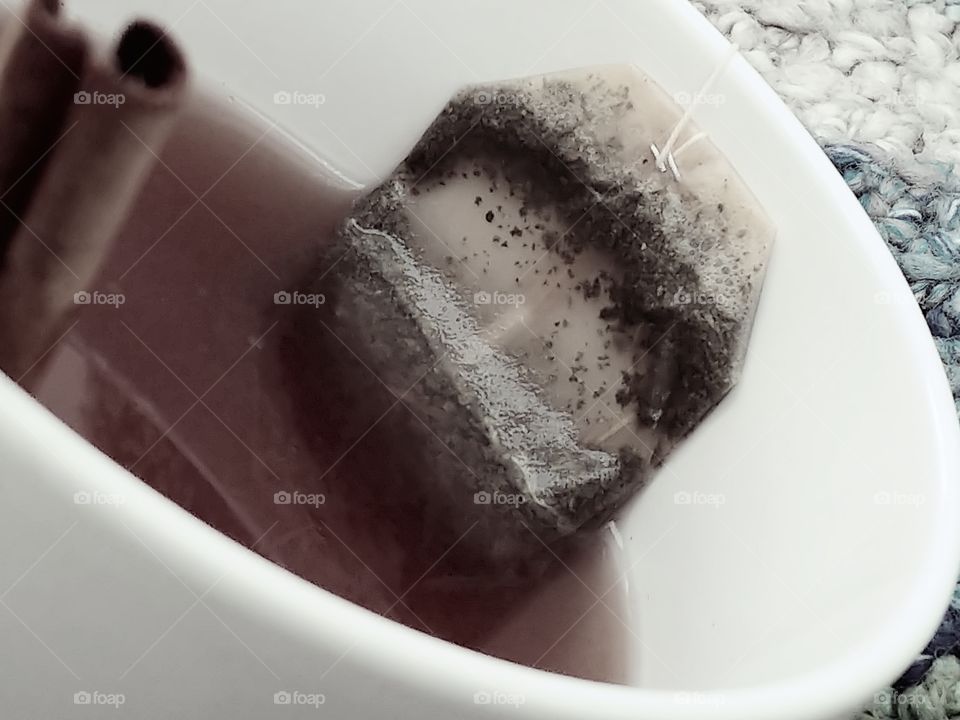 White tea cup filled with dark brown tea with tea bag and cinnamon stick on the side.