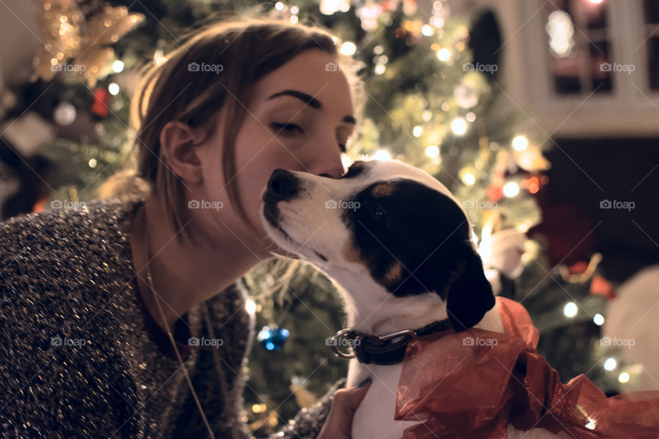 Woman kissing dog with illuminated Christmas tree with red bow on dog 