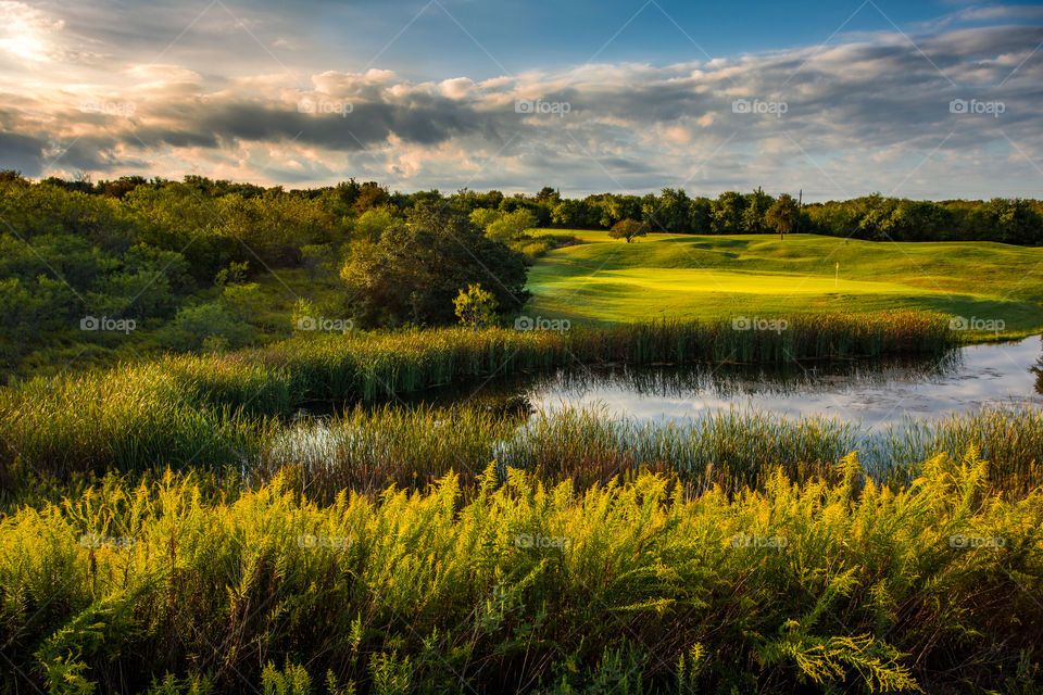 A lush green golf course bathed in the golden glow of the morning sun.  The green is glowing as the warm sunlight peeks out from behind the low morning clouds.  If you look closely, you can see 2 well placed shots next to the flag.
