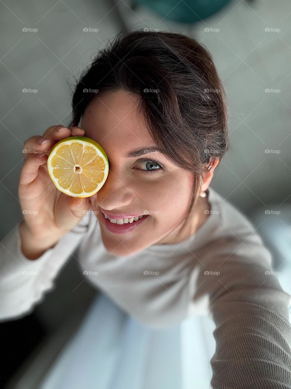 The image shows a woman smiling and holding a slice of lemon over one of her eyes. She is wearing a light gray sweater, and the photo is taken from a top-down angle. The background is softly blurred.