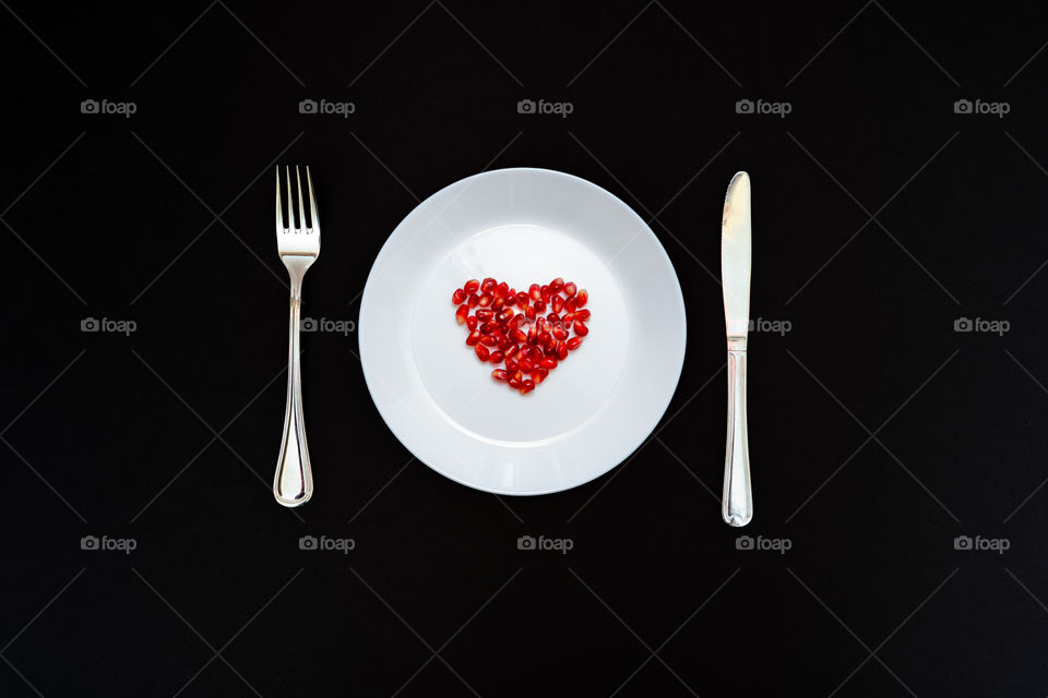 On a white plate from pomegranate seeds composite heart shape. Knife and fork next to the plate. Isolated on black background. Flat layer.