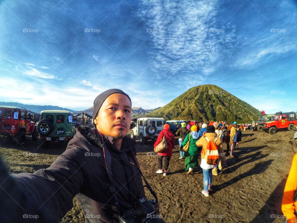 Selfie photo of a young Asian man in warm clothing against a green background in the Bromo mountains area with a cloudy sky. Vacation and adventure.