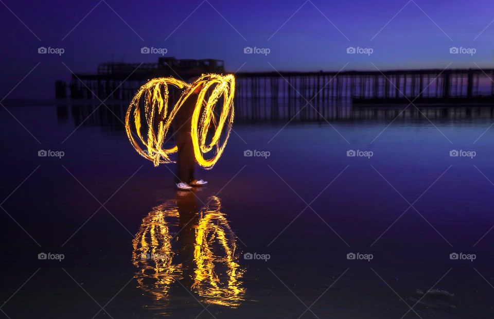 Man spinning fire poi on a wet beach, with an old pier in the background, at dusk